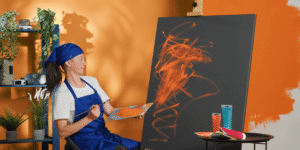 A woman painting on an easel, applying vibrant orange paint to her canvas.
